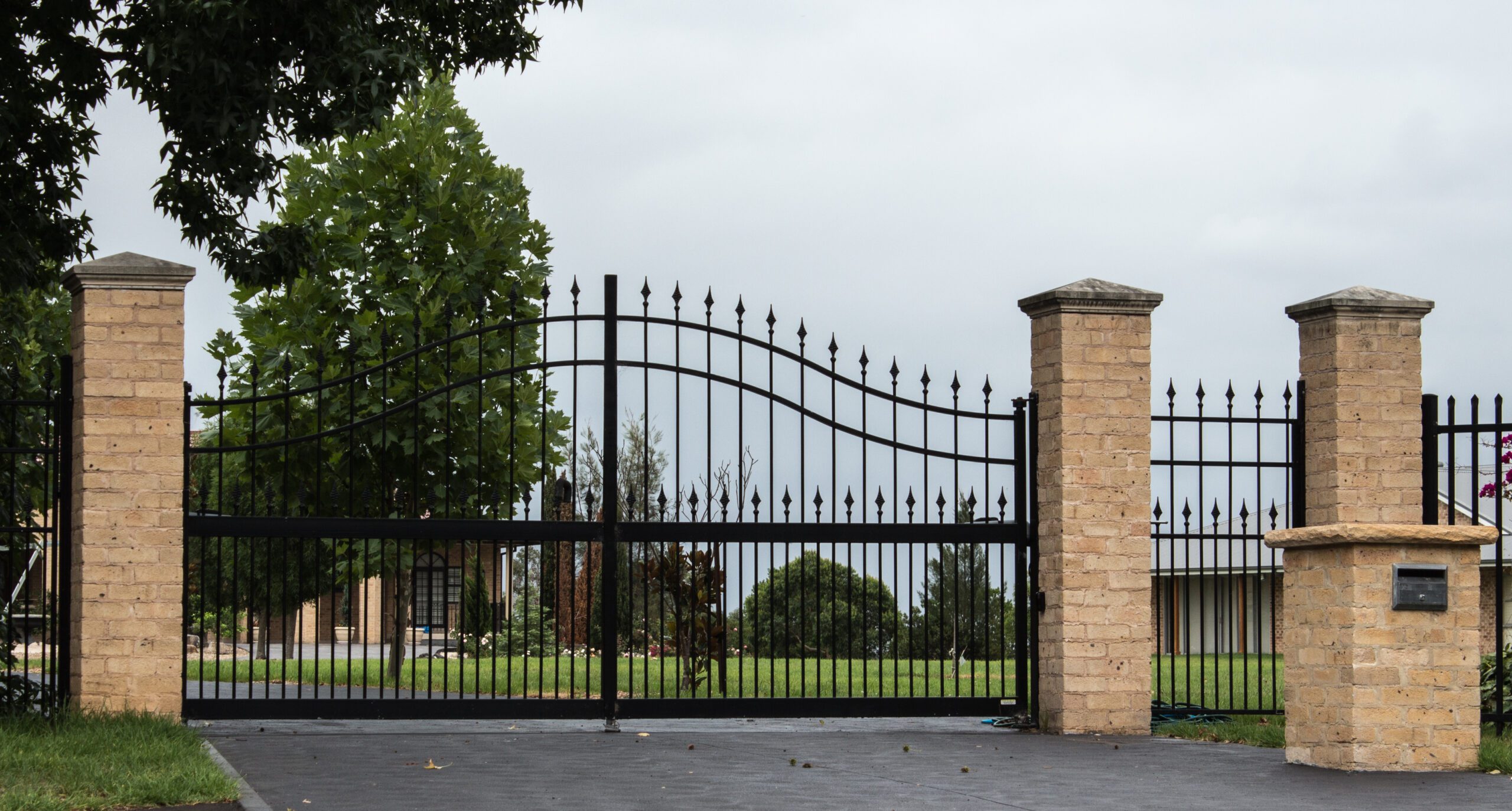 Black metal driveway entrance gates set in brick fence Rénovation et menuiserie à Levignac